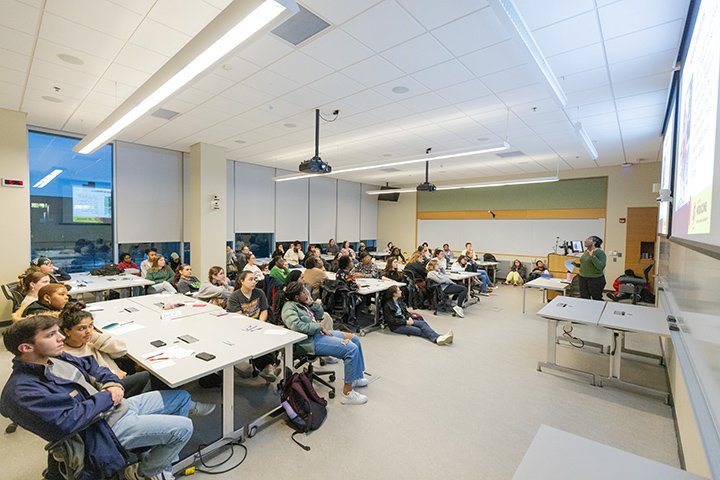 Dozens of students sit at desks in a classroom looking toward the front of the room where a professor speaks.