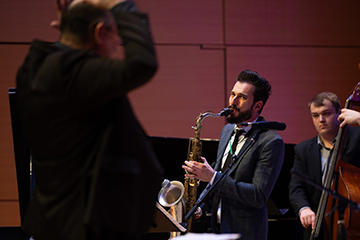 On stage in an auditorium, a saxophone player and  double bass player look towards their conductor in the foreground.