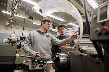 Two students wearing safety glasses talk while standing in a workshop, working on heavy machinery.