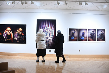 Two women wearing winter coats stand in a brightly lit room with white walls and light brown wood floors. On the walls are large photographs by artist Khary Mason.