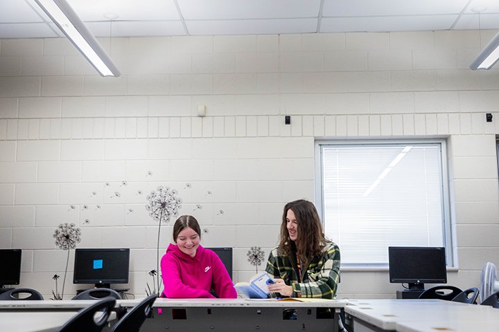 A male CMU student with long hair and wearing a green plaid shirt sits at a desk inside a classroom next to a female high school student in a pink Nike sweatshirt.
