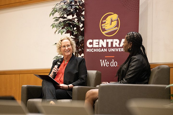 Provost Nancy Mathews sits in a chair holding a microphone, talking to alumna Angle Bush. Behind them is a maroon sign reading