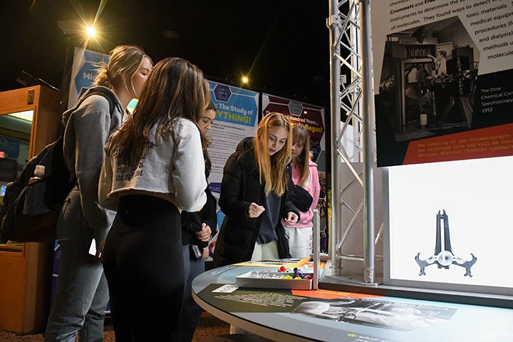 A small group of students stand around the Black Pioneers in Medicine exhibit inside the Rowe Hall. The exhibit is bright and metallic, with displays showcasing contributions of Black individuals in medicine.