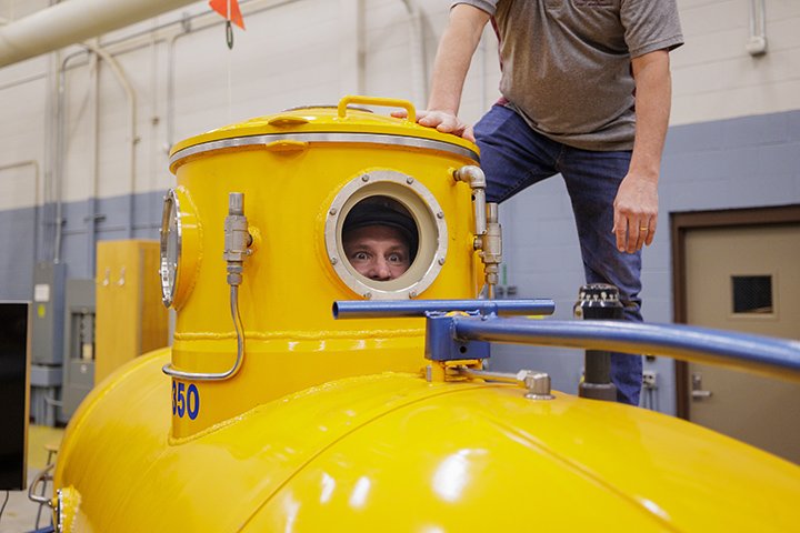 A man peeks through a the window of a small yellow submarine that sits inside a large room.