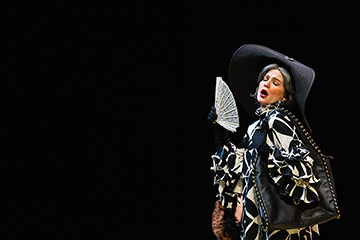 A woman wearing a big black hat and a black and white gown waves a white fan to blow air against her face against a black background.