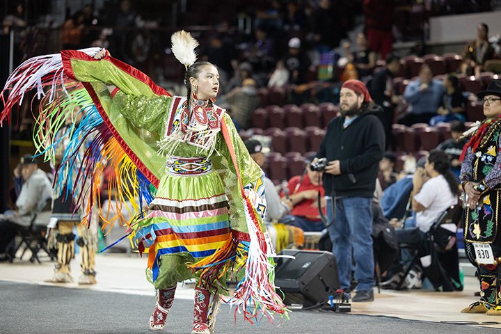 A young woman wearing brightly colored clothing dances on the floor of a gymnasium in front of a crowd.