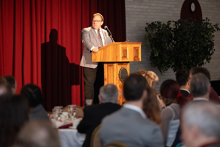 A man stands at a podium in front of a red curtain in front of an audience of people wearing business attire.