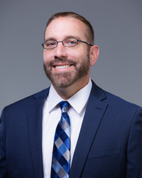 Professional headshot of Shay Dawson, smiling against a gray/blue background while wearing a suit and blue tie.