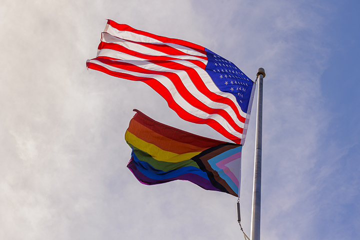 The U.S. flag and Progress Pride Flag wave from a flagpole as thin, white clouds float in the sky.