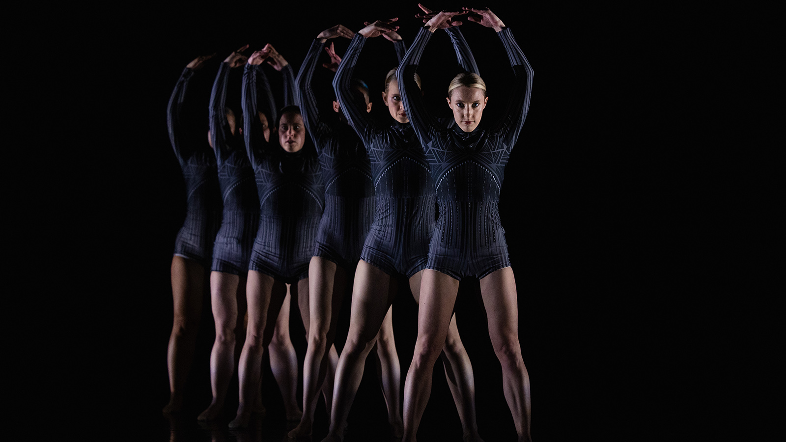 Inside a photo studio with a black backdrop, six CMU dance students wearing dark purple outfits stand in a staggered line with their arms above their heads in the shape of the letter O.