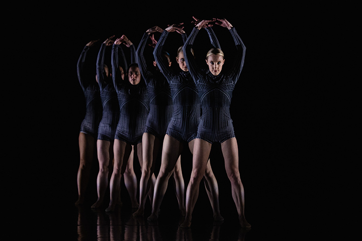 Inside a photo studio with a black backdrop, six CMU dance students wearing dark purple outfits stand in a staggered line with their arms above their heads in the shape of the letter O.