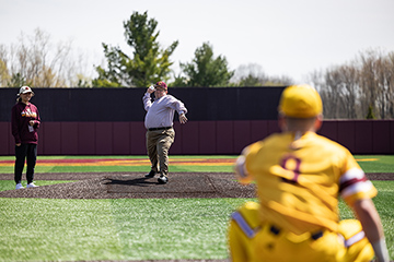 CMU President Bob Davies stands on the mound in the baseball stadium throwing a ceremonial first pitch to a CMU baseball player who squats like a catcher at home plate.