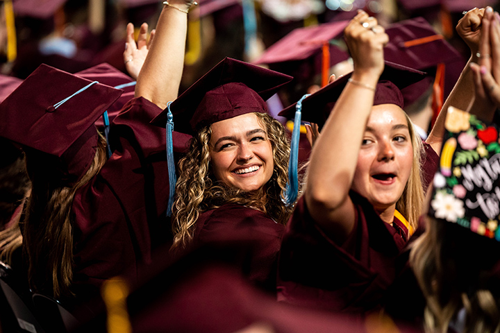 At CMU spring commencement, two graduates with blonde hair sit in chairs, smiling at the crowd and raising their arms.