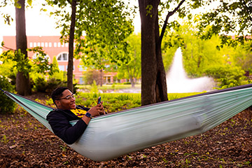 A college student relaxes in a hammock under shaded trees near a pond.