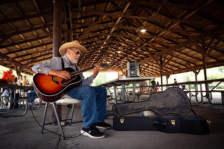 An elderly man wearing sunglasses and blue jeans plays a guitar while sits under a pavilion.