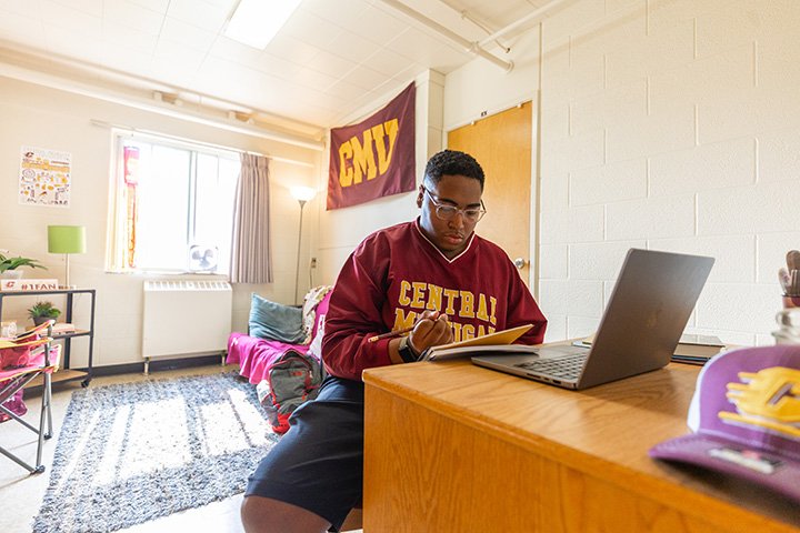 A college student sits at a desk inside a dorm room. On the desk sit a laptop and a CMU hat.