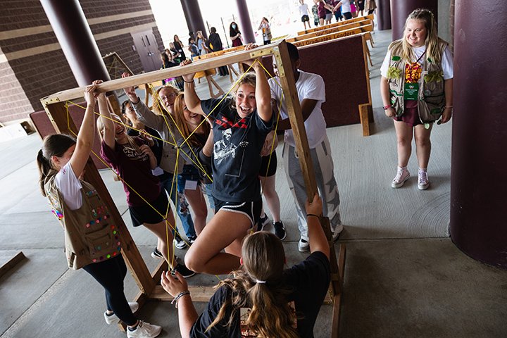 A young woman navigates through a string maze in a wooden frame held by seven other people.