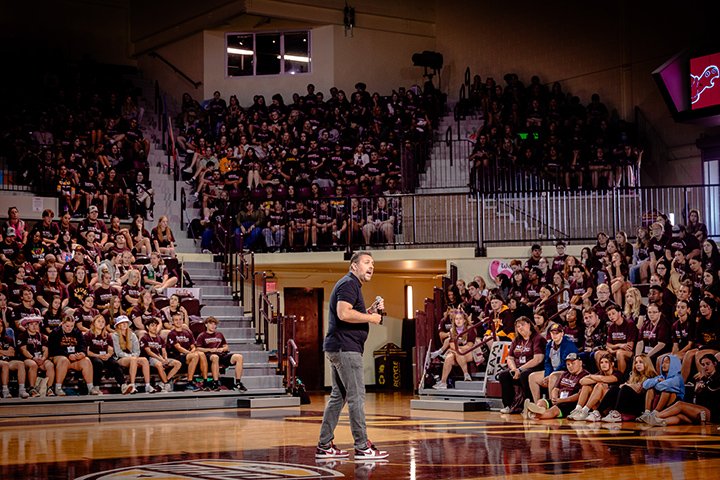 A man in gray pants and a polo shirt holding a microphone stands in the middle of a basketball arena filled with people.
