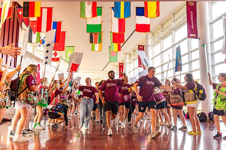 Young people in maroon T-shirts run through a tunnel of other young people beneath flags in a hallway with windows for walls.