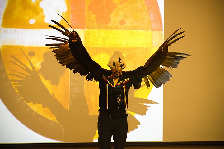 Ty Defoe wears an eagle costume, standing on stage in front of a white and yellow background.