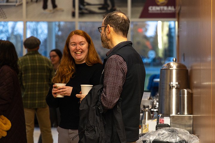 A man and woman stand in an atrium holding coffee and talking.