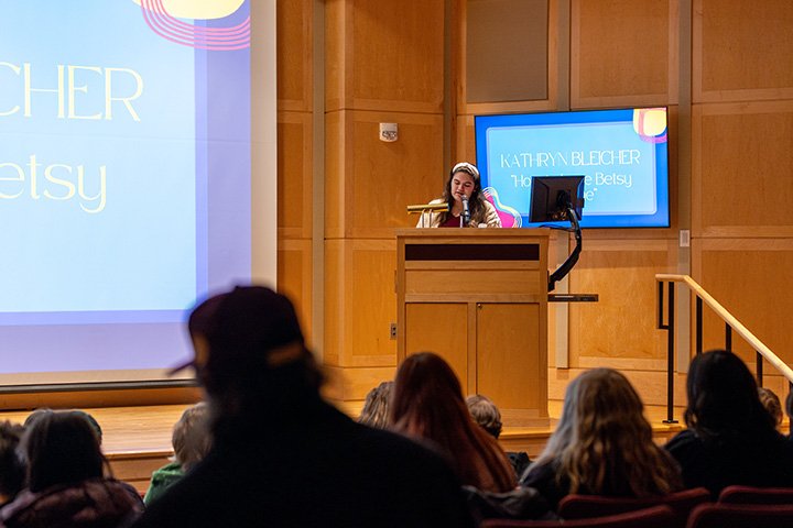 In an auditorium, a young woman stands at a podium giving a presentation.