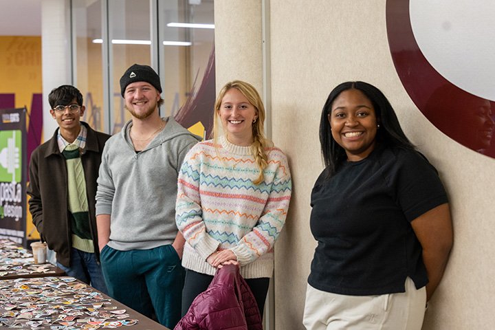 Four students pose for a photo while standing next to a table that has hundreds of stickers sitting on top of it.