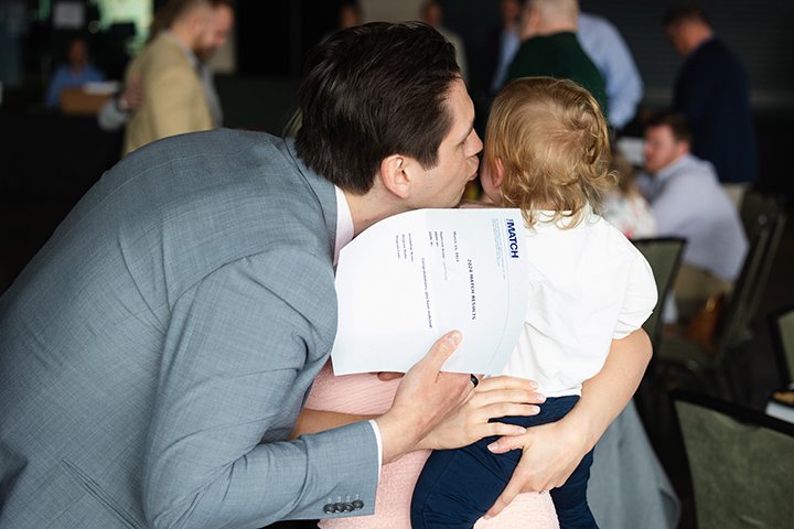 A medical student wearing a gray suit kisses his small child on the cheek after learning where he will do his residency.