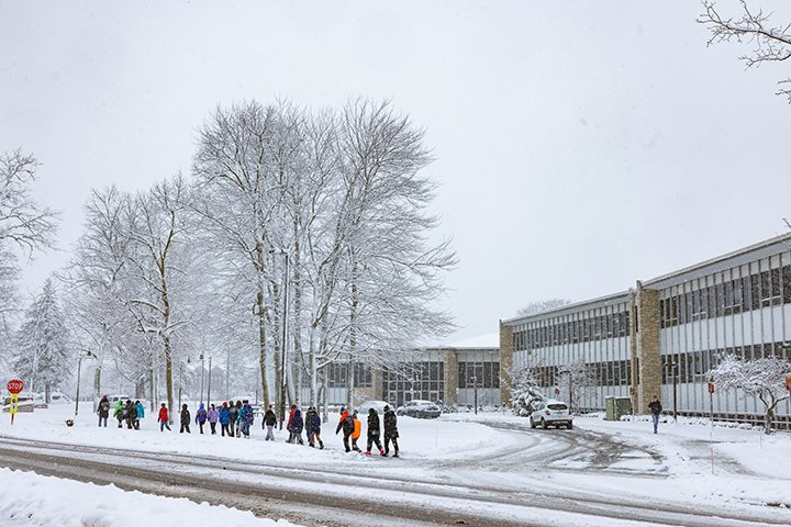 A group of approximately a dozen school-aged students walk along a snow-covered sidewalk in front of the University Center.