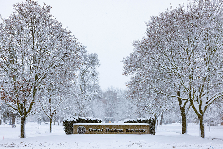 A Central Michigan University sign on the north end of campus stands between two rows of trees with a snowy Warriner Hall in the distance.