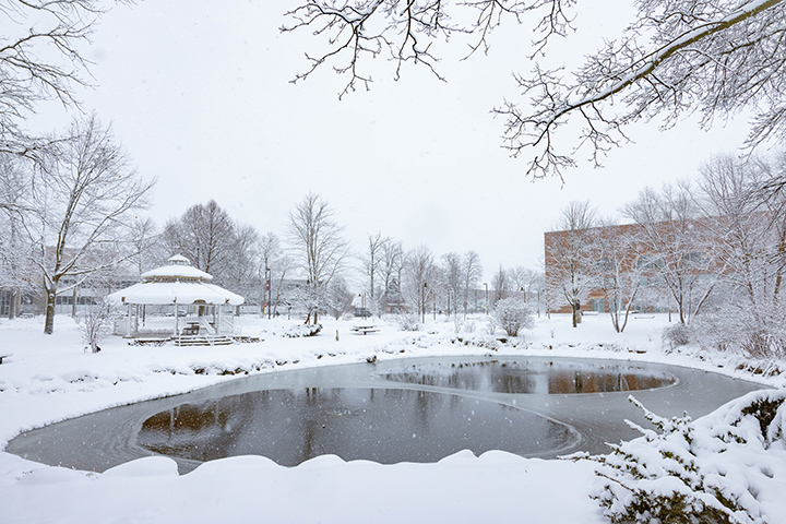 A small ice-covered pond is the focal point of a snowy day image with a gazebo and the University Center in the background.
