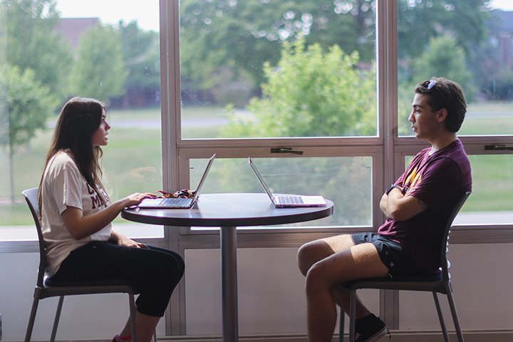 Two college students sit across from each other at a small round table in front of a bright window. Two laptops sit open on the table.