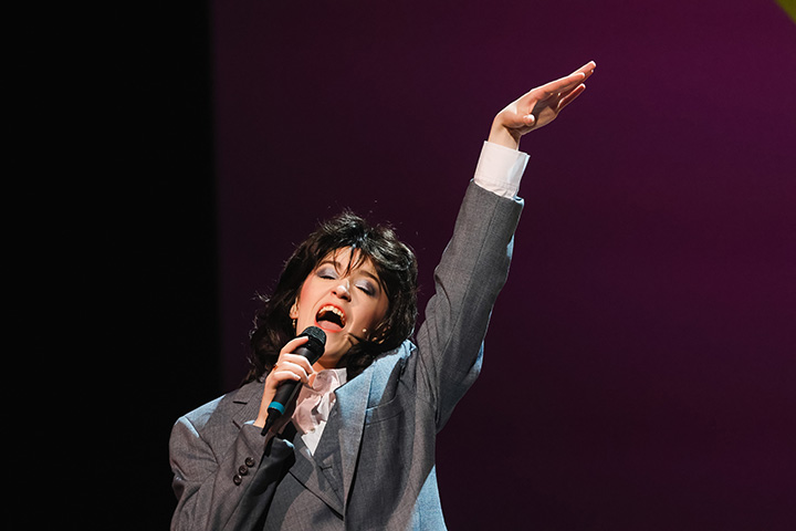 A college student dresses as Pat Benatar while singing on stage during a musical.