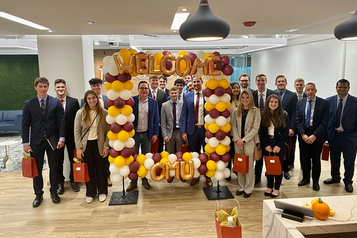 A large group of people wearing suits stand beneath and around a maroon and gold balloon arch.