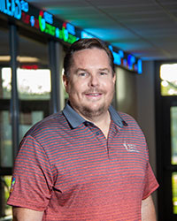 A man with brown hair and a goatee wears a maroon CMU polo shirt.