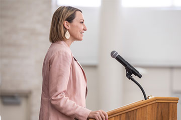 A blond woman in a pink blazer speaks into a microphone.