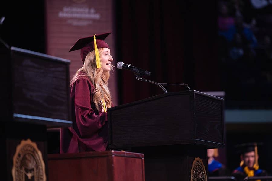 a woman in a maroon graduation cap and gown speaking into a microphone