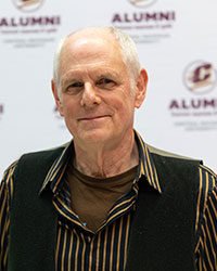 Close-up photo of a smiling man wearing a gold shirt and black vest.