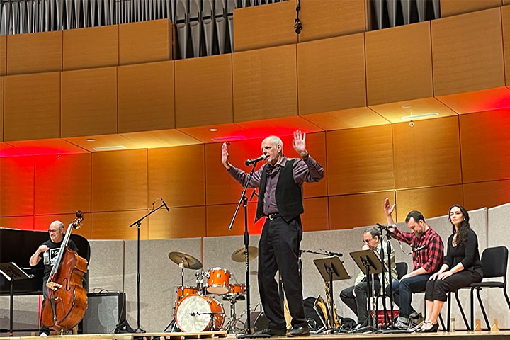 A man stands onstage in front of a microphone while members of a band sit with their instruments around him.