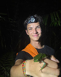 A young man wearing a head lamp holds a green tree frog on his hand.