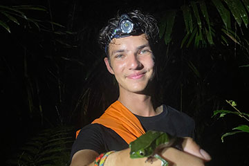 A young man wearing a head lamp holds a green tree frog on his hand.