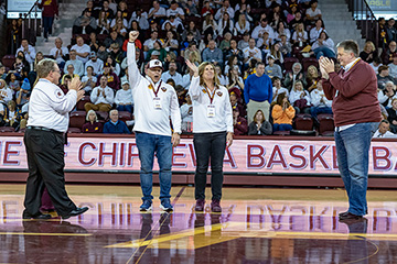 Tim and Sherry Magnusson stand on the basketball court inside McGuirk Arena raising their hands as the crowd cheers.