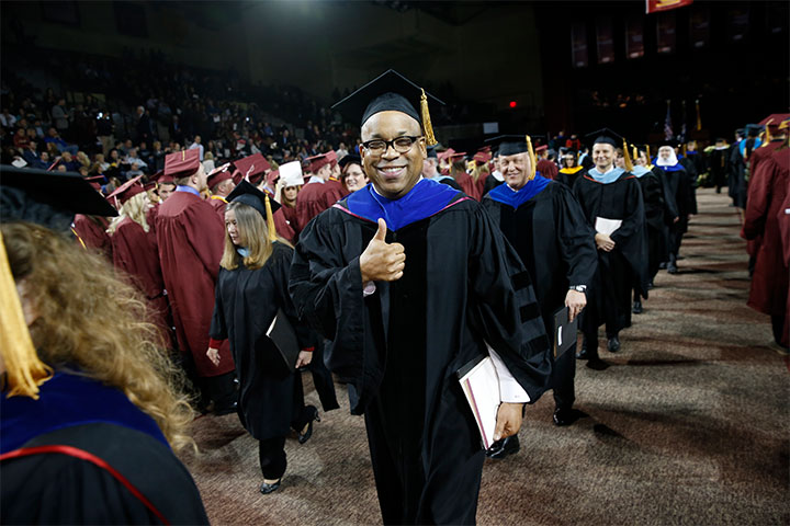 A man wearing a graduation cap and gown smiles at the camera and gives a thumbs up.