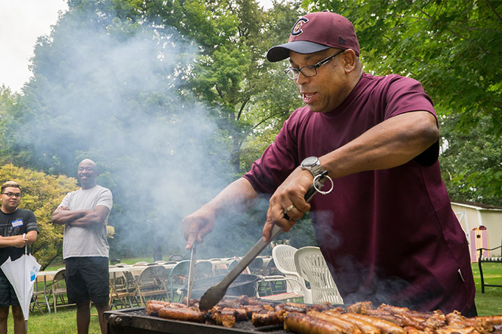 A man in a maroon shirt and hat cooks hot dogs on a barbeque grill.
