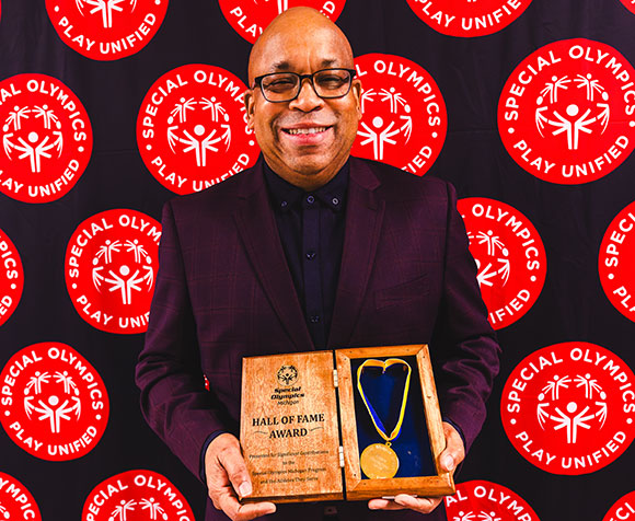 A Black man poses in front of a Special Olympics backdrop holding an award.