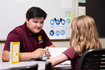 A male teacher wearing a maroon shirt talks to a little girl holding a notebook in a classroom setting.