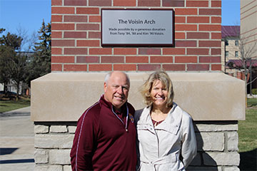 A man in a maroon sweater stands to the left of a woman in a white jacket, both appear in front of a plaque that reads The Voisin Arch.