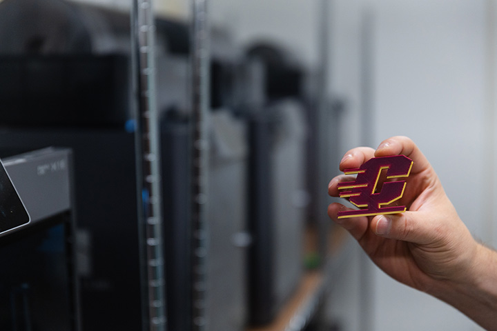 A hand holding a small 3D-printed Action C in the foreground with 3D printers blurred in the background