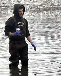 A man with a sweatshirt hood pulled up over his head and wearing safety gloves holds a sample jar while standing in water.