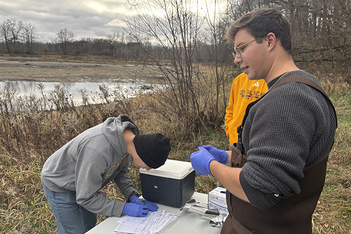 Two young men wearing blue safety gloves process water samples outside on a plastic table.